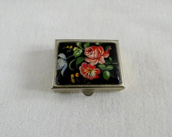 May include: A small, silver-coloured metal box with a hinged lid. The lid features a floral design with red, pink, and yellow flowers on a black background.