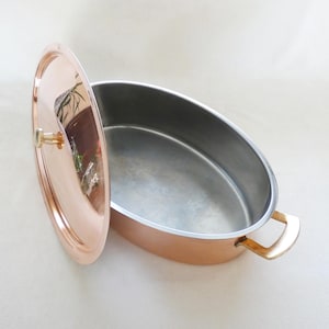 May include: An oval copper roasting pan with a polished exterior and a silver-toned interior lining. The pan features two brass-coloured side handles and comes with a matching circular copper lid that has a small brass-coloured knob handle.
