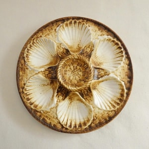 May include: A round, ceramic oyster serving platter with a brown and beige color scheme. The platter features six shell-shaped compartments and a central well, all with a textured design. The outer rim has a woven basket pattern.