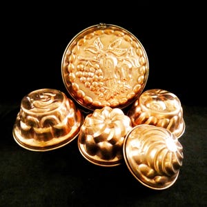 May include: A collection of copper molds, including a large round one with a grape and leaf design, and several smaller, decorative molds. The molds have a shiny, reflective surface and are arranged against a dark background.