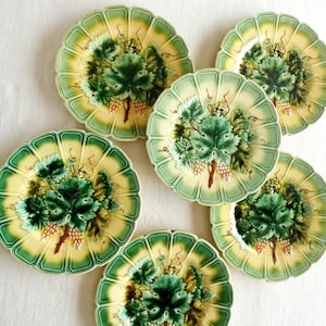 May include: Six decorative plates with a scalloped edge. Each plate features a central design of green grape leaves and a cluster of grapes, set against a yellow and green background. The plates are arranged on a white surface.