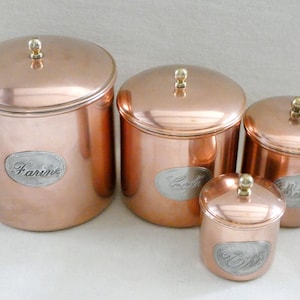 Puede incluir: Juego de cuatro botes de cocina de cobre con tapas y etiquetas plateadas. Las etiquetas dicen "Farine", "Cookies", "Biscuits" y "Té". Cada bote tiene un pomo dorado. Los botes son de diferentes tamaños.