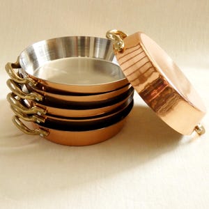 Puede incluir: Una pila de pequeñas sartenes de cobre con asas de latón y un interior brillante y reflectante. Una sartén descansa de lado, mostrando el exterior de cobre. Estas sartenes son probablemente para cocinar y añaden un toque de elegancia a cualquier cocina.