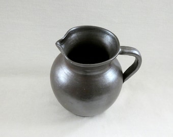 May include: A dark brown ceramic pitcher with a rounded body and a single handle. The pitcher has a smooth, glossy finish.