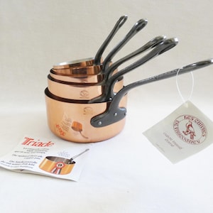 May include: A set of copper saucepans with black handles, stacked together. The pans have a shiny, reflective surface. A tag with text and a brochure with a picture of a saucepan are also present.