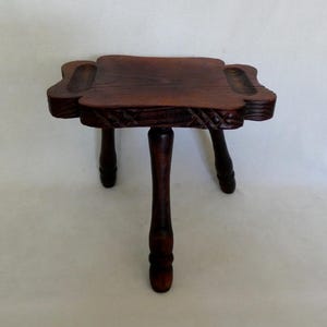 May include: A small, dark brown wooden stool with three turned legs. The square seat features rounded corners with carved indentations on each side and a decorative cross-hatched pattern along the edges of the top surface, showcasing a visible wood grain.
