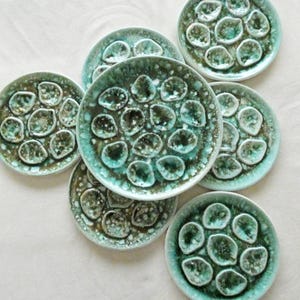 Puede incluir: Un conjunto de siete platos de cerámica con un esmalte moteado turquesa y marrón. Cada plato presenta un patrón de formas redondeadas en relieve, creando una superficie texturizada. Los platos están dispuestos sobre una superficie blanca.