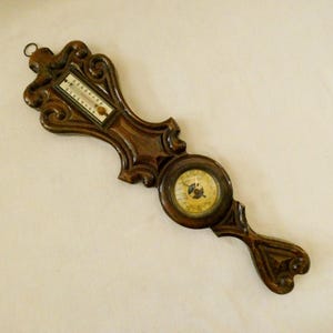 May include: A dark brown wooden wall-mounted weather station featuring a vertical thermometer with a white scale and a circular barometer with a cream-colored dial. The wood frame has ornate, carved scrollwork details and a metal hanging loop.