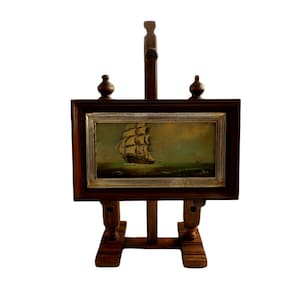 Può includere: Un dipinto rettangolare di un veliero sull'oceano, incorniciato in argento e legno scuro, montato su un cavalletto in legno marrone scuro con due terminali decorativi sulla parte superiore e un supporto centrale con un pomello rotondo.