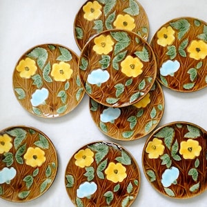 Puede incluir: Un juego de ocho platos redondos de cerámica con un fondo marrón de veta de madera, hojas verdes en relieve y flores amarillas y azul claro. Cada plato muestra un patrón floral consistente en toda la superficie con un acabado brillante.