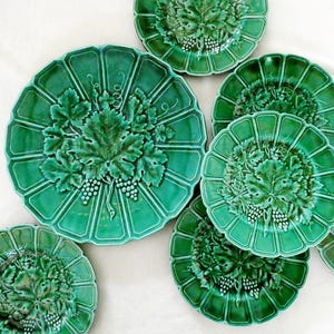 May include: A collection of seven green ceramic plates featuring a raised relief design of grape leaves and clusters of grapes in the center. Each plate has a scalloped edge and a glossy finish, arranged in a cluster on a plain white surface.