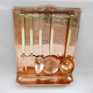 May include: Copper kitchen utensil rack with five hanging tools. The rack has a rectangular shape with a curved top and a small shelf at the bottom. The utensils include a slotted spoon, ladle, and spatula.