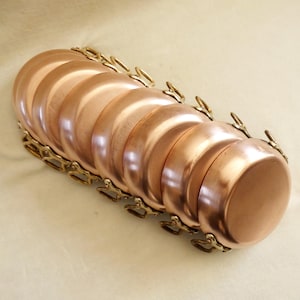May include: A collection of ten small copper saucepans, each with brass handles and a decorative brass chain. The pans are arranged in a row, displaying their shiny surfaces. The copper color is a warm, inviting tone.
