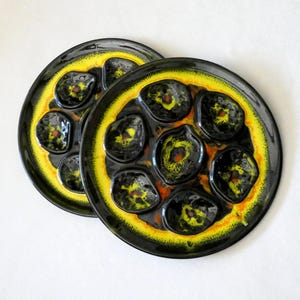 Może przedstawiać: Dwa czarne ceramiczne talerze do ślimaków z żółtymi i pomarańczowymi akcentami. Każdy talerz ma siedem oddzielnych przegródek. Talerze mają błyszczące wykończenie i są ustawione na białym tle.