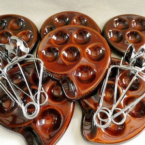 Pode incluir: Conjunto de seis ramequins de cerâmica marrom com alças, cada um com seis reentrâncias individuais. Os ramequins estão empilhados com um conjunto de pinças de metal.