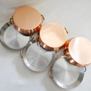 May include: A collection of copper and stainless steel pans. The copper pans have a shiny, reflective surface and brass handles. The stainless steel pans have a brushed metal finish and brass handles. The pans are round and appear to be for cooking.