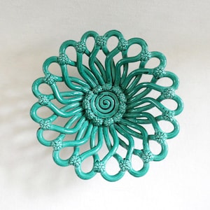 May include: A circular teal ceramic bowl featuring an intricate openwork design. The center displays a spiral pattern surrounded by small flower-shaped accents. Radiating outward are curved, looped clay strands that form a scalloped edge.