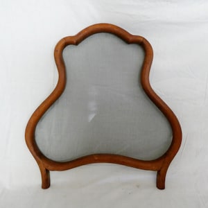 Può includere: Un paravento in legno per camino con una cornice curva e ornata. La cornice è di colore marrone caldo e circonda uno schermo a rete grigia. Lo schermo ha una forma decorativa unica e poggia su due piccole gambe.