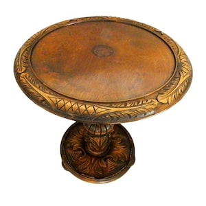 May include: A vintage, ornate wooden side table with a round top and a detailed, carved base. The table is made of a warm, brown wood with decorative carvings around the edge of the tabletop and on the pedestal base. The table is approximately 61 cm tall.