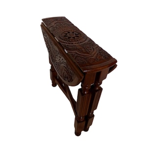 May include: A dark brown wooden folding table featuring intricate floral and geometric carvings on the top surface and side panels. The table has four turned legs and is shown in a collapsed, upright position with its side leaves folded down.