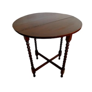 Op de afbeelding: Een ronde, donkerbruine houten tafel met een gespleten blad. De tafel heeft vier gedraaide poten met een kruisvormige steun. De tafel heeft een diameter van ongeveer 76 cm.