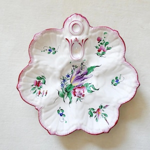 Może przedstawiać: Biały ceramiczny półmisek z falistym brzegiem, ręcznie malowany we wzory kwiatowe w kolorze różowym, fioletowym i zielonym. Obrzeże jest obramowane ciemnoczerwoną obwódką. Półmisek ma uchwyt i centralne wgłębienie.