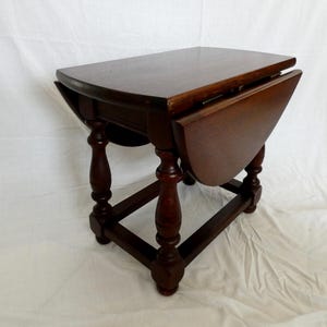 May include: A dark brown wooden drop-leaf table featuring a rectangular top with rounded edges and two hinged side leaves. The table is supported by four turned legs connected by a rectangular base frame, showcasing a classic furniture design.