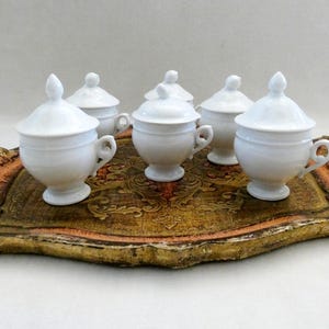 May include: A set of five white porcelain lidded cups with handles and pedestal bases, arranged on a rectangular decorative tray featuring a distressed gold and brown finish with an intricate embossed pattern across the surface.