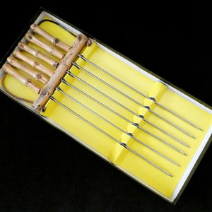 May include: A set of six metal skewers with wooden handles. The skewers are in a yellow box with a white border. The handles are made of natural bamboo.