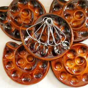 May include: Five brown ceramic escargot serving dishes, each with multiple indentations for holding snails. A set of silver-toned snail tongs rests on one dish. The dishes have a glossy finish.