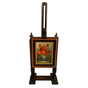 May include: A dark brown wooden easel holding a rectangular framed painting of orange and red flowers in a vase. The easel features a vertical adjustable support post, a sturdy base, and decorative finials on the top corners of the frame holder.