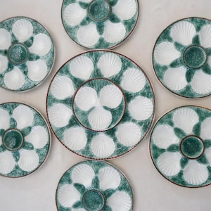 Może przedstawiać: Zestaw ceramicznych talerzy do ostryg. Każdy talerz ma biały wzór muszli na turkusowo-zielonym tle. Talerze są ułożone w okrąg, podkreślając ich dekoracyjny charakter.