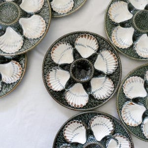 May include: A collection of round ceramic oyster plates featuring a dark green, textured basket-weave border and a central circular well. Each plate contains six white, shell-shaped indentations with brown-tinted edges arranged in a radial pattern.