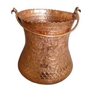May include: A copper-colored metal bucket with a handle. The bucket has an embossed floral design and a scalloped rim. The handle is attached to the rim with decorative accents. The bucket has a wide base and tapers inward.