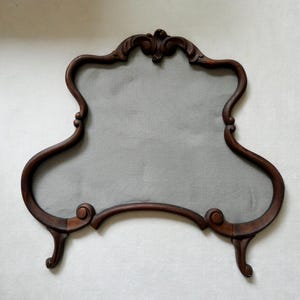 May include: A decorative fireplace screen featuring a dark brown wooden frame with ornate, curved edges and carved scroll details at the top and base. The center panel is filled with a solid, light gray fabric insert, creating a contrast.