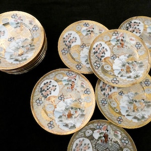 Op de afbeelding: Een set ronde keramische borden met een goudkleurige rand en een centraal ontwerp van waaiervormige motieven, bloempatronen en kleine figuren. De borden hebben een witte basis met gedetailleerde gouden, zwarte en oranje accenten.