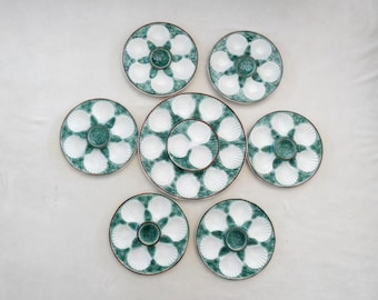 May include: A set of ceramic oyster plates featuring a white seashell design on a teal backdrop. The plates are arranged in a circular pattern, with a larger plate at the centre and smaller plates around it. The plates have a brown rim.