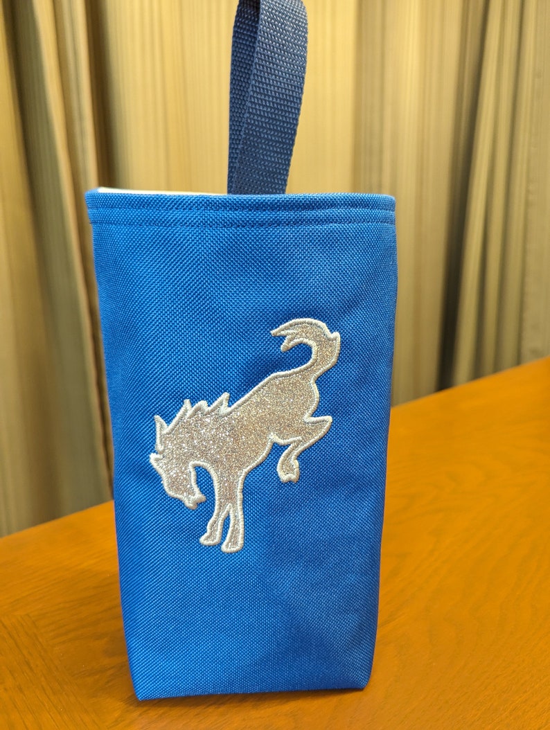 May include: A blue canvas tote bag with a silver glitter embroidered horse design. The bag has a navy blue handle.