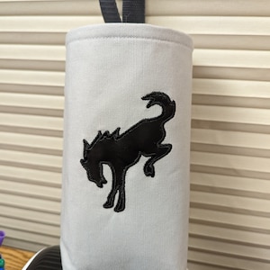 May include: A white canvas bottle holder with a black embroidered running horse design. The holder has a grey fabric loop handle.