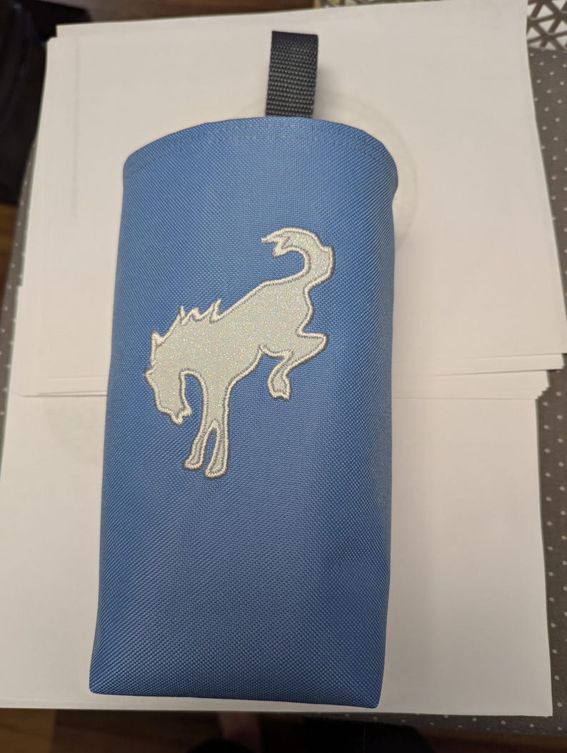 May include: A blue fabric pouch with a white embroidered horse design. The pouch has a black strap at the top.