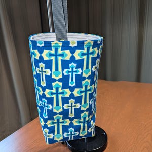 May include: A blue and green fabric holder with a cross pattern for a paper towel roll. The holder is attached to a black stand with a gray strap.