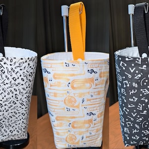 May include: Three fabric cup holders with different music-themed patterns. The first cup holder has a black and white pattern of musical notes. The second cup holder has a white and yellow pattern of musical notes and brass instruments. The third cup holder has a black and white pattern of musical notes.