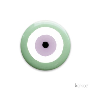 May include: A round, painted eye-shaped design with a light green outer ring, a white inner ring, a lavender center, and a black pupil. The design is centered on a white background. The word "kökoa" is in the bottom right corner.