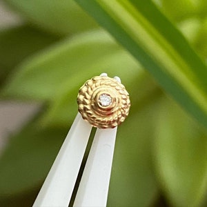 Peut inclure: Boucle d'oreille à clou dorée avec un motif circulaire texturé et une petite pierre précieuse transparente au centre. La boucle d'oreille est tenue par une pince à épiler blanche sur un fond vert flou.