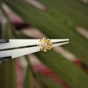 May include: A gold stud earring with a central clear gemstone, held by white tweezers. The earring features a sunburst design with a beaded border. The background is a blurred mix of green and pink.