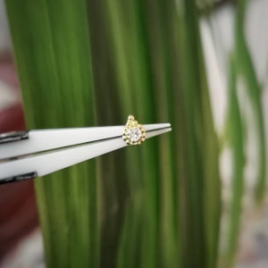 Peut inclure: Une petite boucle d'oreille en or avec une pierre précieuse ronde et transparente au centre. La boucle d'oreille est tenue par une pince à épiler blanche sur un fond vert flou, mettant en valeur son design délicat et ses détails scintillants.