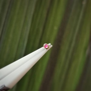 May include: A tiny, gold-coloured stud earring featuring a round, pink gemstone, held by white tweezers. The earring is set against a blurred green backdrop, implying a natural setting. The gemstone is the central element.