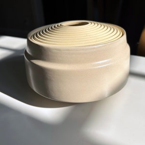 May include: A beige ceramic container with a layered, concentric design on the top. The container has a smooth, matte finish.