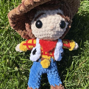 Puede incluir: Mu&ntilde;eca de ganchillo hecha a mano de un personaje de vaquero, con sombrero marr&oacute;n, cara blanca y atuendo rojo, amarillo y azul. La mu&ntilde;eca est&aacute; hecha de hilo suave y texturizado y est&aacute; colocada sobre hierba verde.