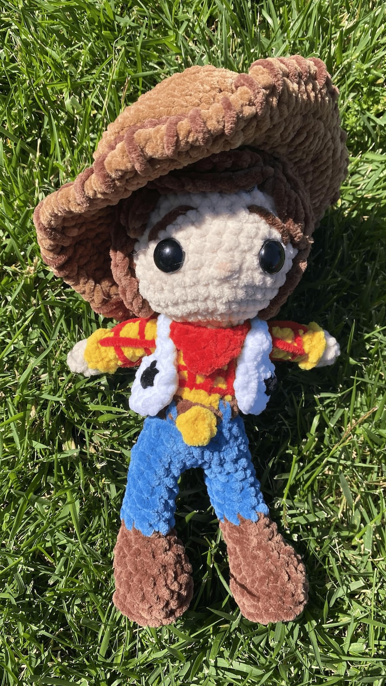 Puede incluir: Mu&ntilde;eca de ganchillo hecha a mano de un personaje de vaquero, con sombrero marr&oacute;n, cara blanca y atuendo rojo, amarillo y azul. La mu&ntilde;eca est&aacute; hecha de hilo suave y texturizado y est&aacute; colocada sobre hierba verde.